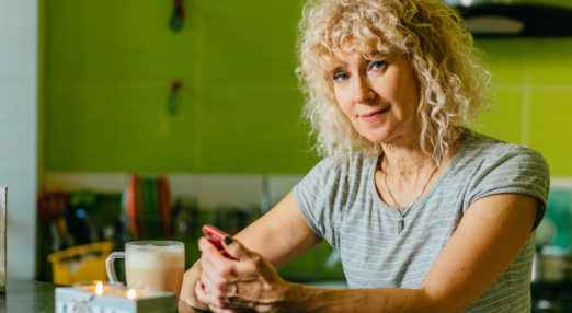 Image of blonde woman sitting at a table with a phone and a cup of coffee looking straight to camera