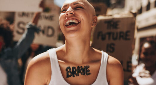 Image of a bald woman with "brave" on her chest at a protest