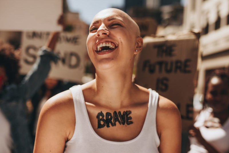 Image of a bald woman with "brave" on her chest at a protest