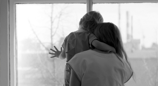 Black and white photo of a women and her child looking out of a window on to an urban winter landscape. The child places their hand on the window as if they want to go outside.