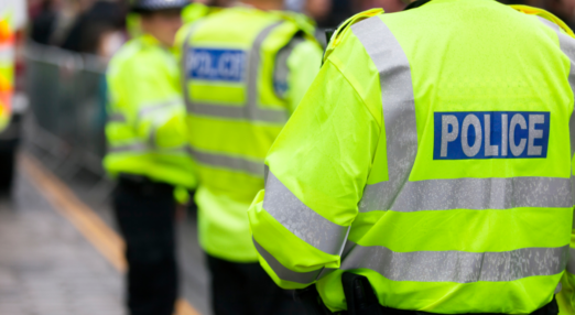 Police Officer from the back in a high-visibility jacket reading 'POLICE'. In the background, two more officers can be seen.