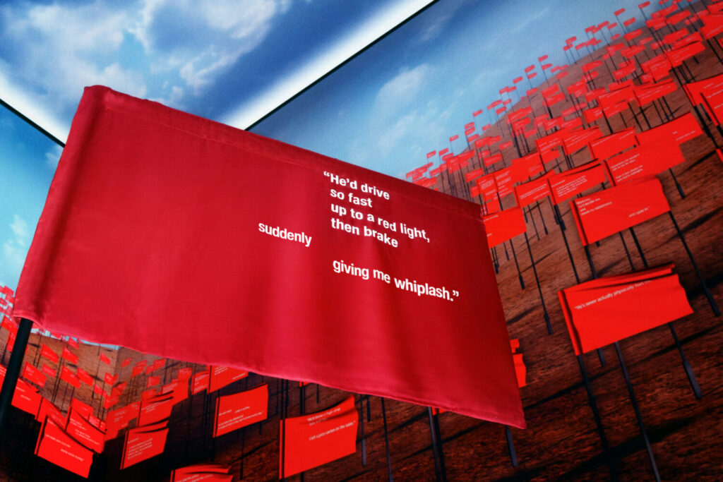 Red flag on a pole reading "He'd drive so fast up to a red light, then brake suddenly giving me whiplash". Behind it is a screen filled with red flags