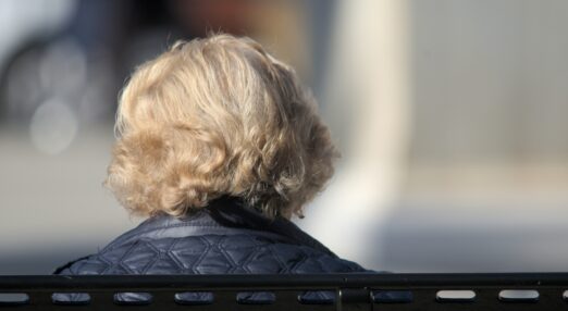 White haired woman photographed from the back sat on a park bench