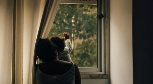A Black mother and her young child sit looking out a window, facing away.