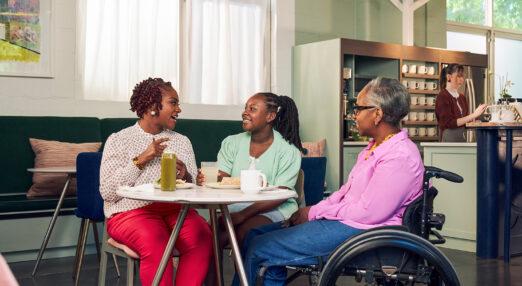 Three Black women sat round a table in a coffee shop. On the left, the woman is signing to the woman in the middle, who has a cochlear implant