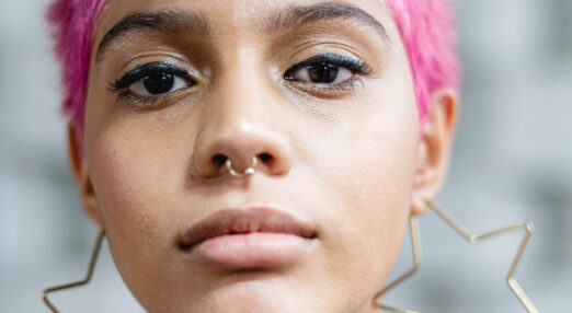 Image of the face of a young non-binary person with short pink hair, looking seriously at the camera.