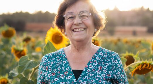 An older, white woman smiles in a field of sunflowers with the sun behind her